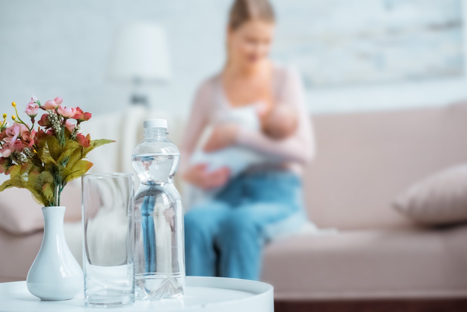Auf einem kleinen Sofatisch stehen eine Vase, ein Wasserglas und eine Plastikflasche Wasser; im unscharfen Hintergrund sitzt eine Frau mit einem Baby im Arm auf dem Sofa