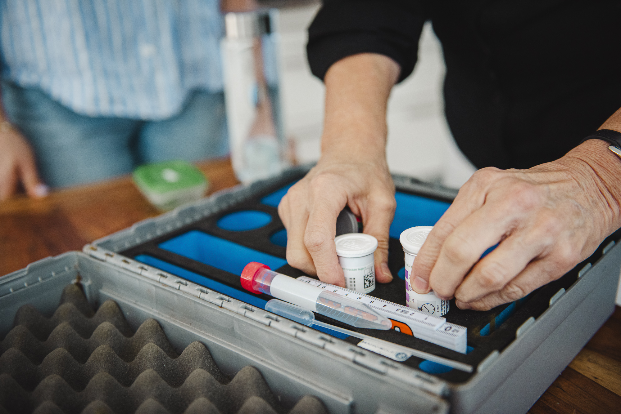 Koffer mit Testzubehör für einen Wassertest liegt auf einem Tisch, Anja Freese greift nach Zubehör, im Hintergrund steht eine Frau am Tisch