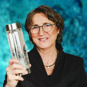 Anja Freese hält eine Wasserflasche mit Sprudelwasser vor einem Hintergrund aus Luftblasen im Wasser